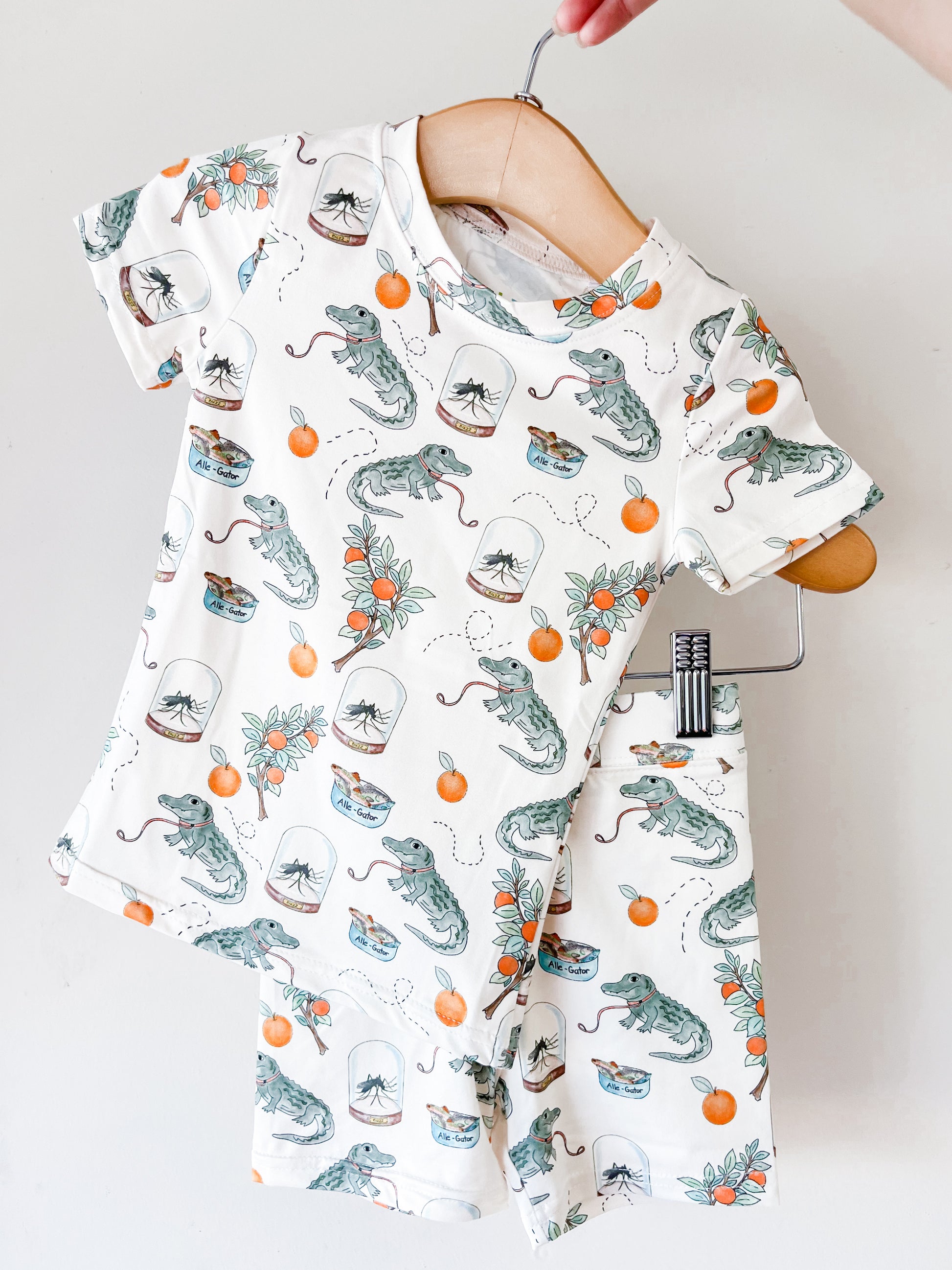 The Tiny Tot Bundles Sunshine Swamp Lounge Set hangs on wooden hangers, showcasing a white short-sleeve shirt and shorts playfully printed with alligators, oranges, orange blossoms, and little blue bowls.