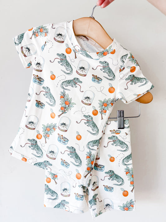 The Tiny Tot Bundles Sunshine Swamp Lounge Set hangs on wooden hangers, showcasing a white short-sleeve shirt and shorts playfully printed with alligators, oranges, orange blossoms, and little blue bowls.