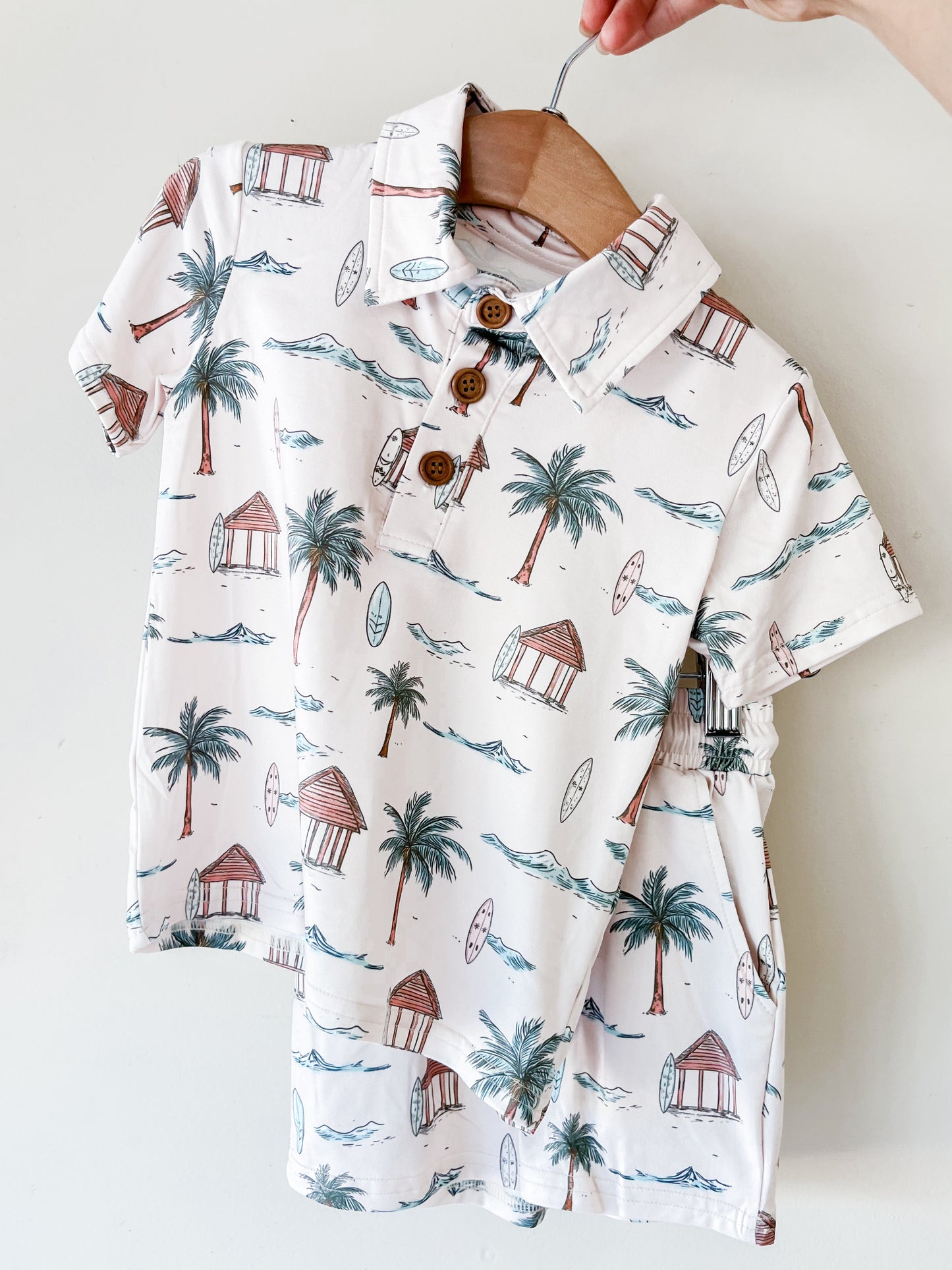 A hand holds the Tiny Tot Bundles Palm Coast Collared Set—a child’s short-sleeved white shirt with a tropical print of palm trees, surfboards, huts, and waves, and three front buttons—displayed on a wooden hanger.