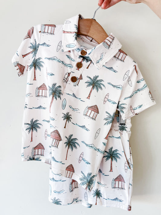 A hand holds the Tiny Tot Bundles Palm Coast Collared Set—a child’s short-sleeved white shirt with a tropical print of palm trees, surfboards, huts, and waves, and three front buttons—displayed on a wooden hanger.