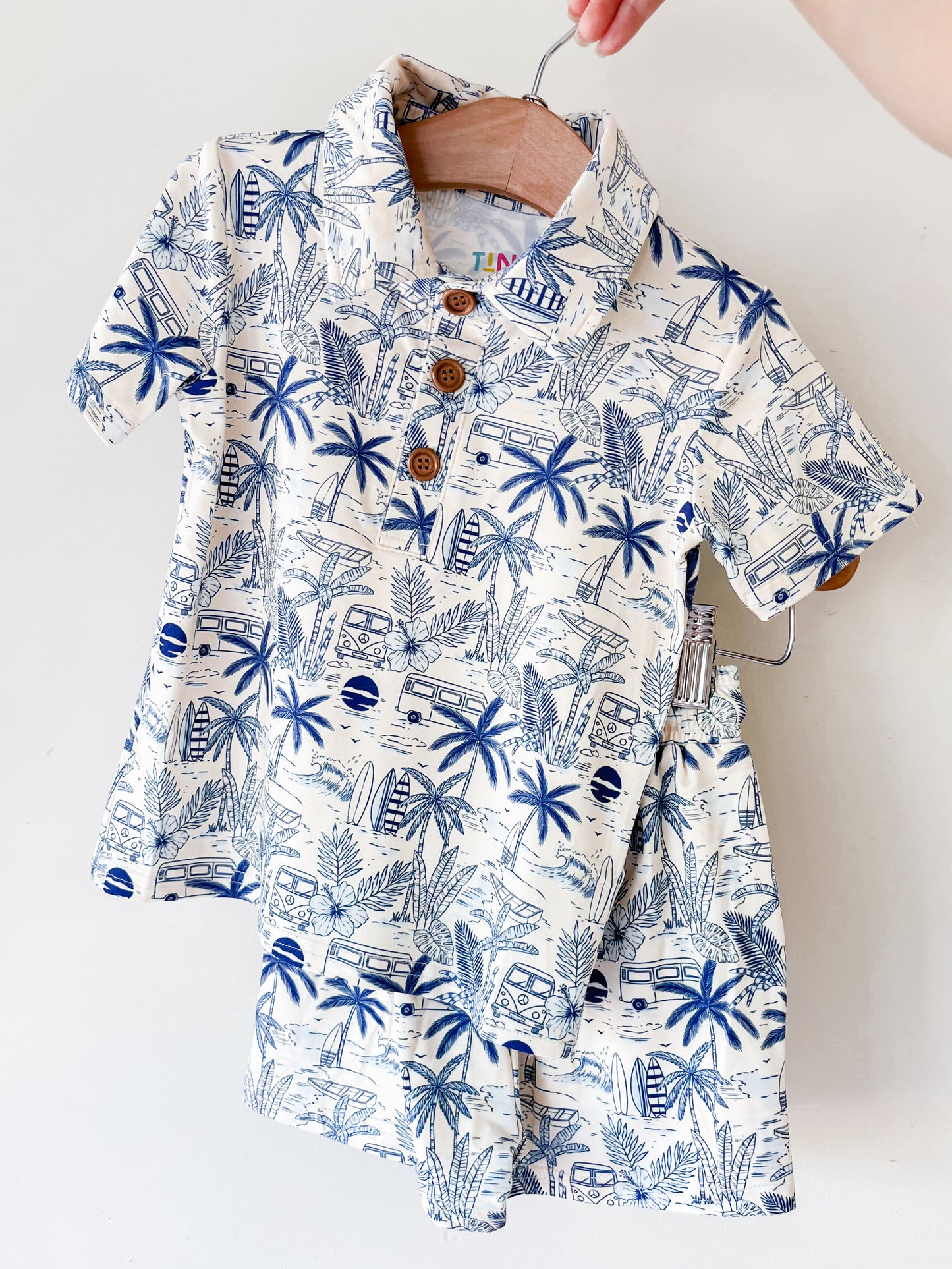 A hand holds up the Surfside Florida Collared Set by Tiny Tot Bundles, a matching children’s outfit with a collared shirt and shorts featuring a blue and white tropical print of palm trees, surfboards, and vans.