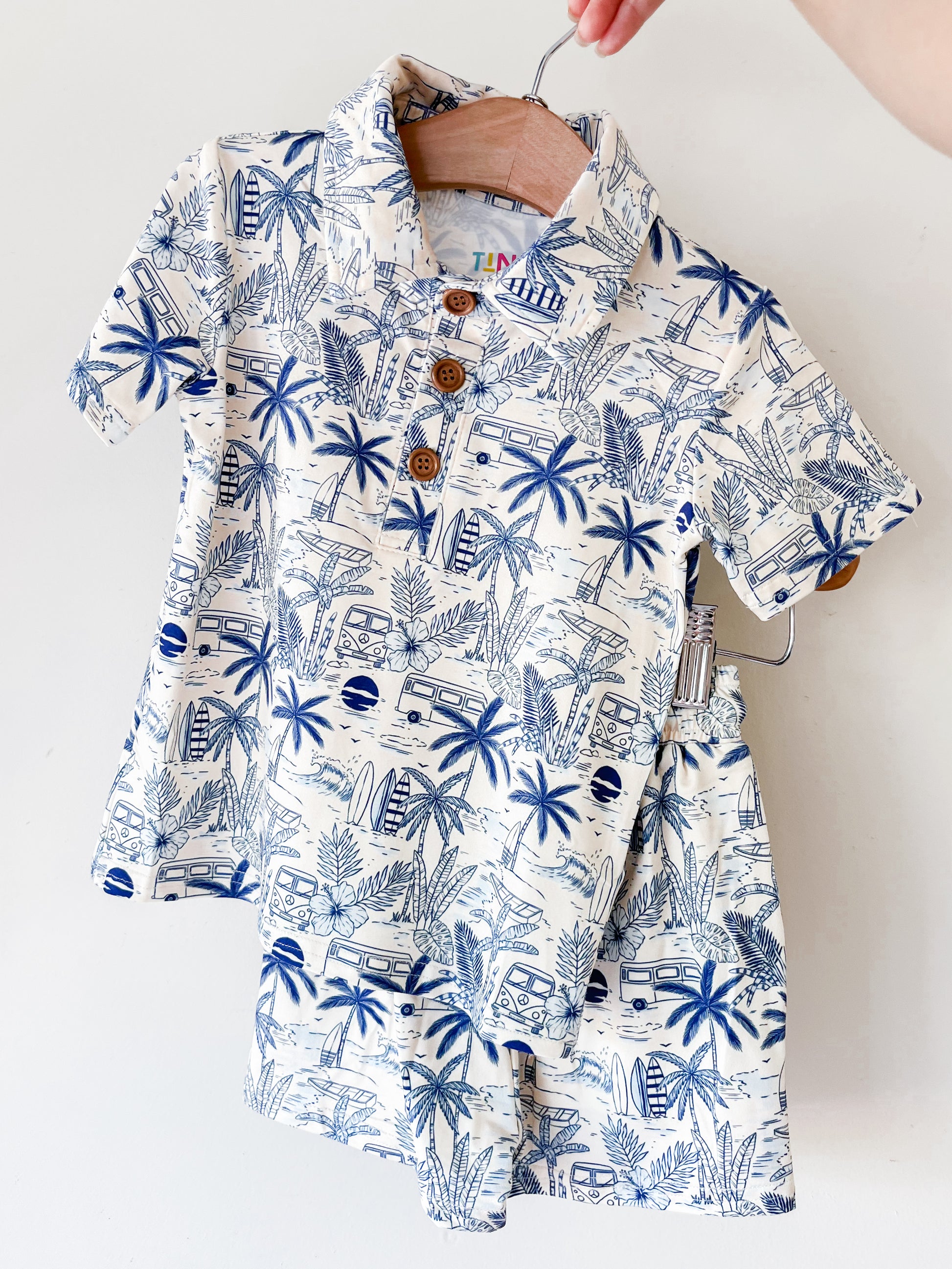 A hand holds up the Surfside Florida Collared Set by Tiny Tot Bundles, a matching children’s outfit with a collared shirt and shorts featuring a blue and white tropical print of palm trees, surfboards, and vans.