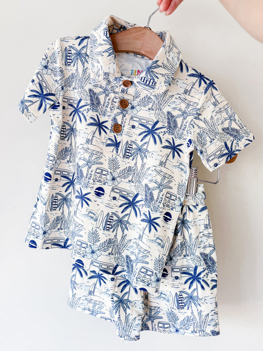 A hand holds up the Surfside Florida Collared Set by Tiny Tot Bundles, a matching children’s outfit with a collared shirt and shorts featuring a blue and white tropical print of palm trees, surfboards, and vans.