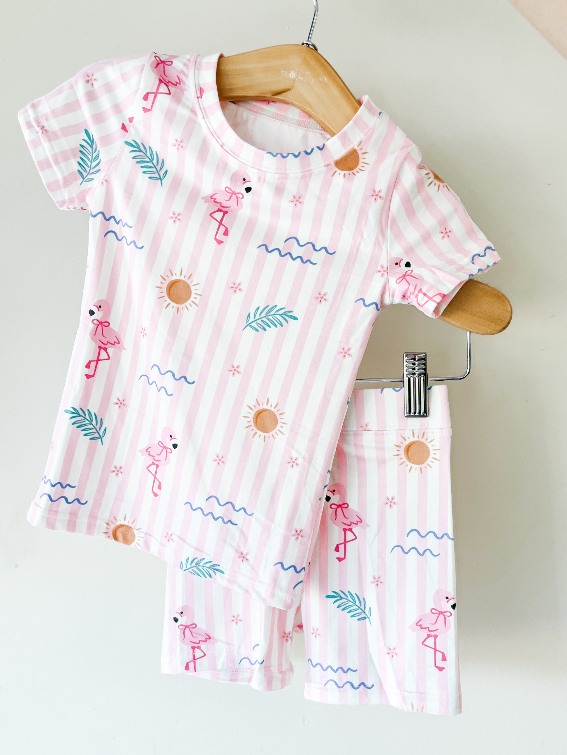 The Pink Paradise Lounge Set by Tiny Tot Bundles is a children’s pink and white striped short-sleeve pajama set with flamingo, sun, leaf, and wavy line patterns, displayed on wooden hangers against a white background.