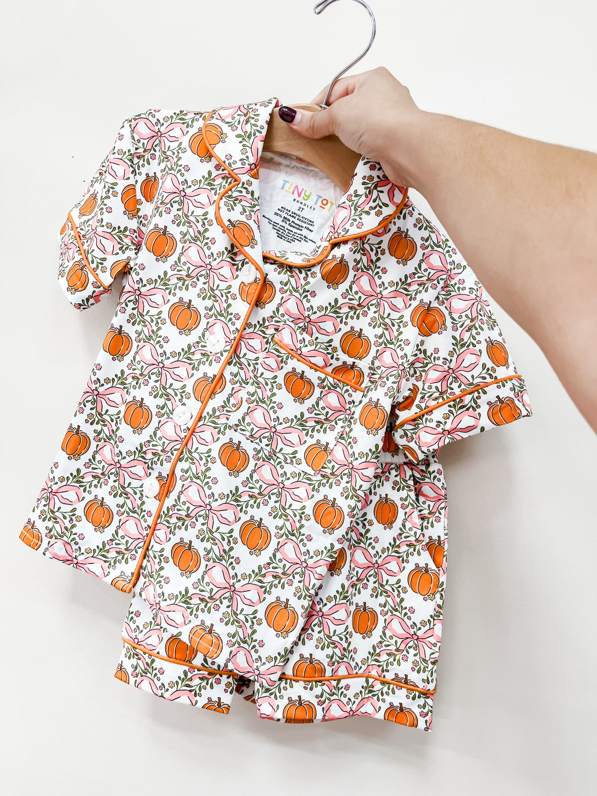A hand holds up a Tiny Tot Bundles Sweet Autumn Short Jammies Set with an orange pumpkin and pink bow pattern on a wooden hanger.