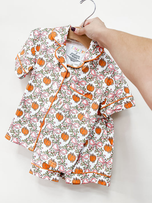 A hand holds up a Tiny Tot Bundles Sweet Autumn Short Jammies Set with an orange pumpkin and pink bow pattern on a wooden hanger.