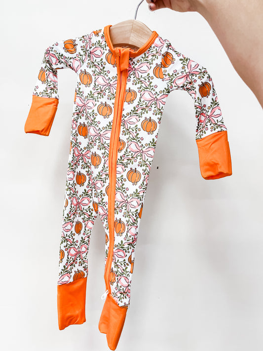 Tiny Tot Bundles Sweet Autumn Zippy long-sleeved baby sleeper with a zipper closure, featuring a print of orange pumpkins and pink bows on a white background with orange cuffs.