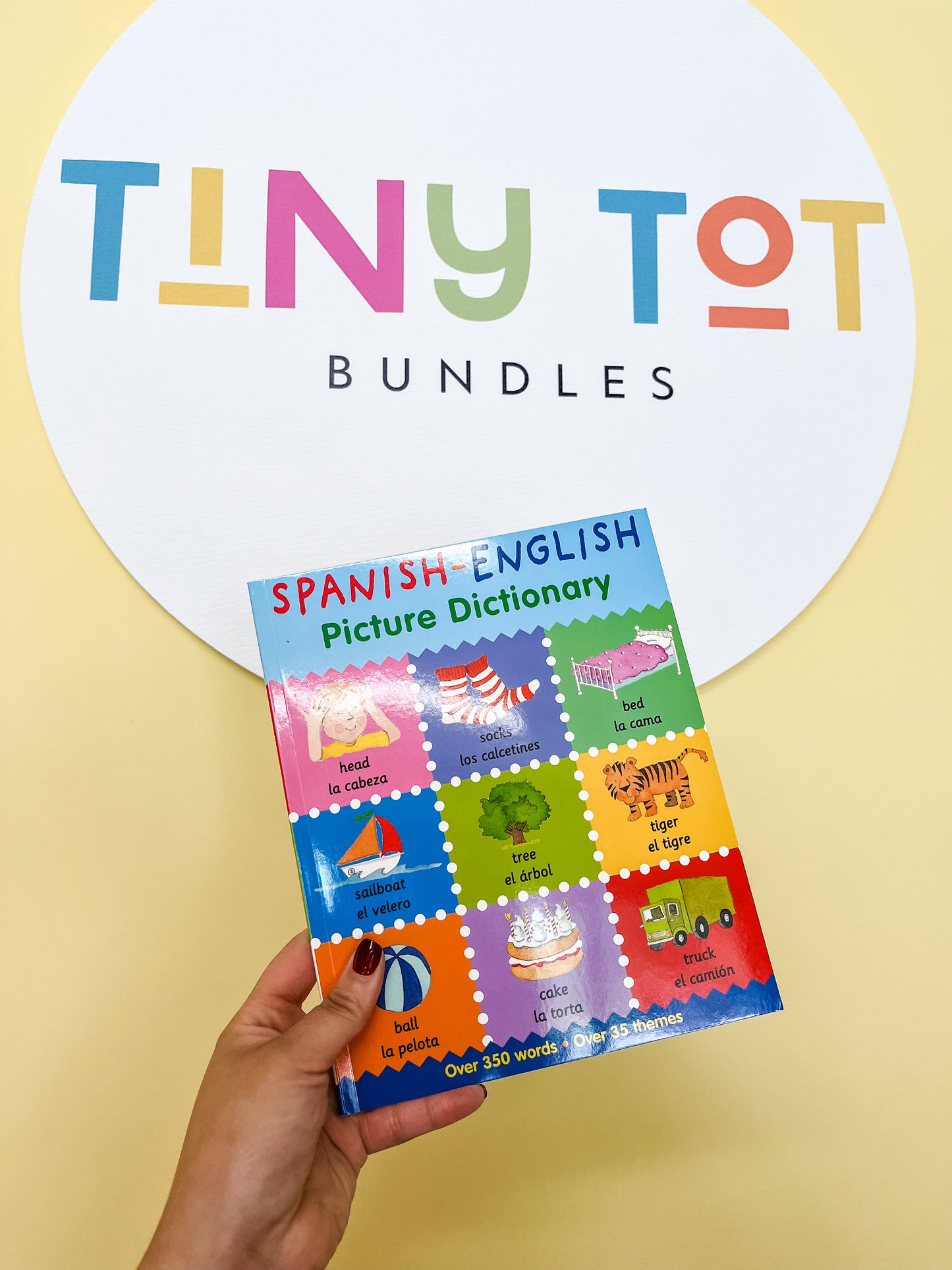 A hand holds the Spanish-English Picture Dictionary Book by Tiny Tot Bundles, featuring bilingual words and colorful illustrations. In the background, a round sign displays the brand name on a yellow wall.