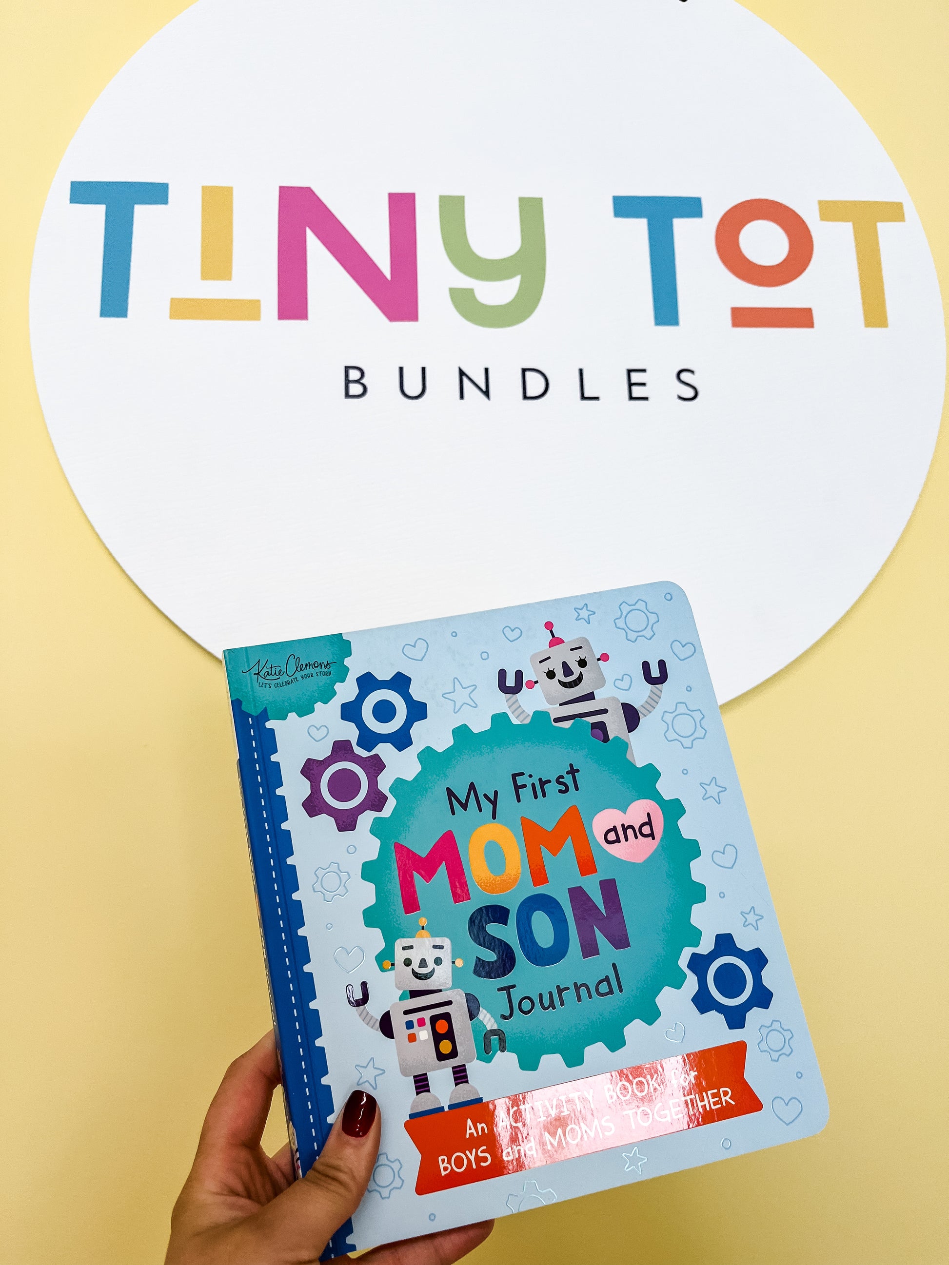 A hand holds the colorful My First Mom & Son Journal by Tiny Tot Bundles in front of a round sign with the brand's playful, multicolored logo.