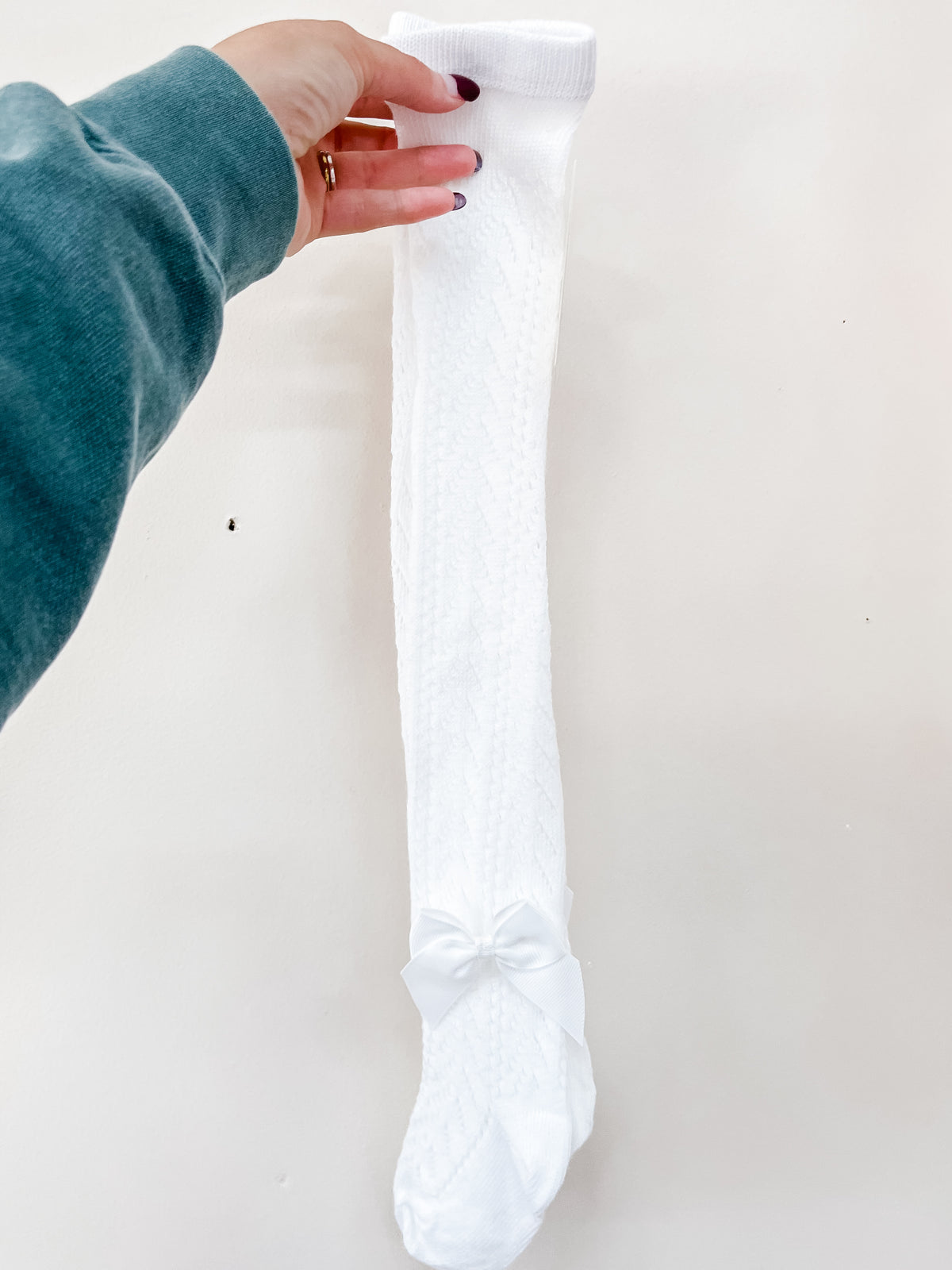A hand holds up a white, knee-high Sweet Bow Detail Knit Socks pair featuring a small ribbon bow at the ankle.