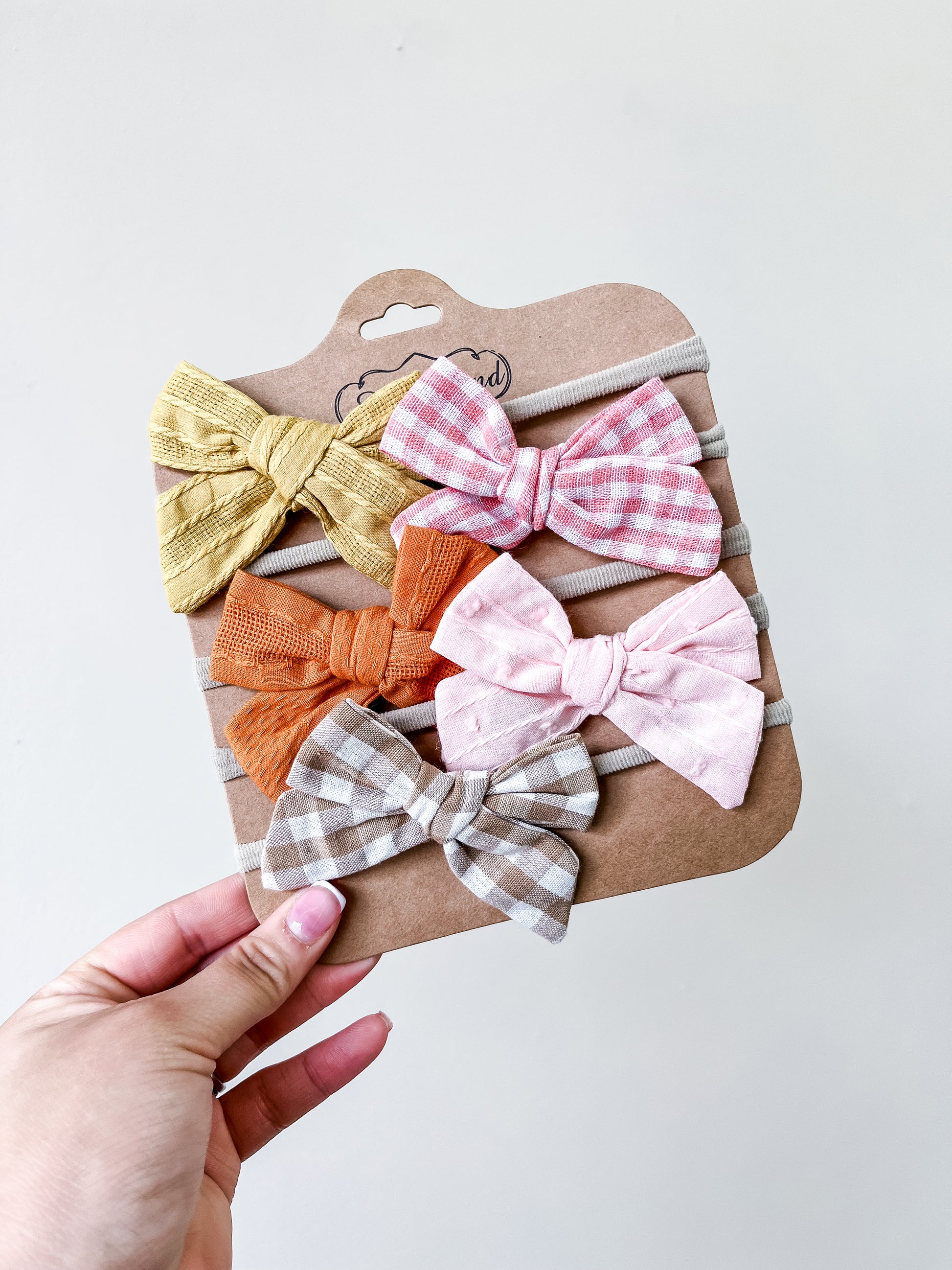 A hand holds the Bloom Bow Set by Tiny Tot Bundles, showing four baby headbands with yellow, orange, pink plaid, and gray plaid bows arranged in a row against a plain light background.