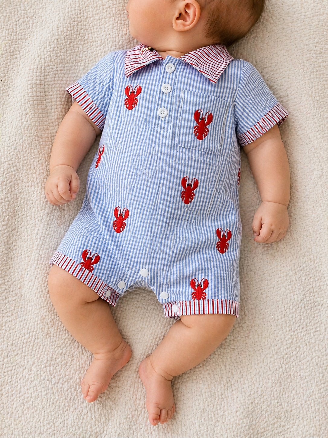 A baby in the Tiny Tot Bundles Lobster Dock Romper, featuring blue and white stripes with red lobsters and red-striped trim, lies relaxed on a soft cream blanket, head turned to the side.