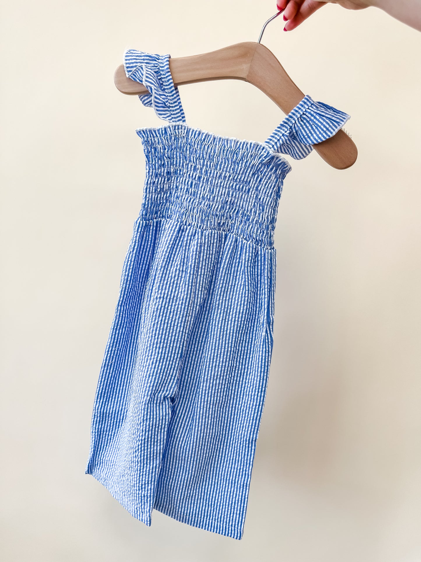 A hand holds the Blue Stripe Smocked Jumpsuit by Tiny Tot Bundles—a sleeveless, ruffled romper with blue and white stripes—displayed against a plain light background.