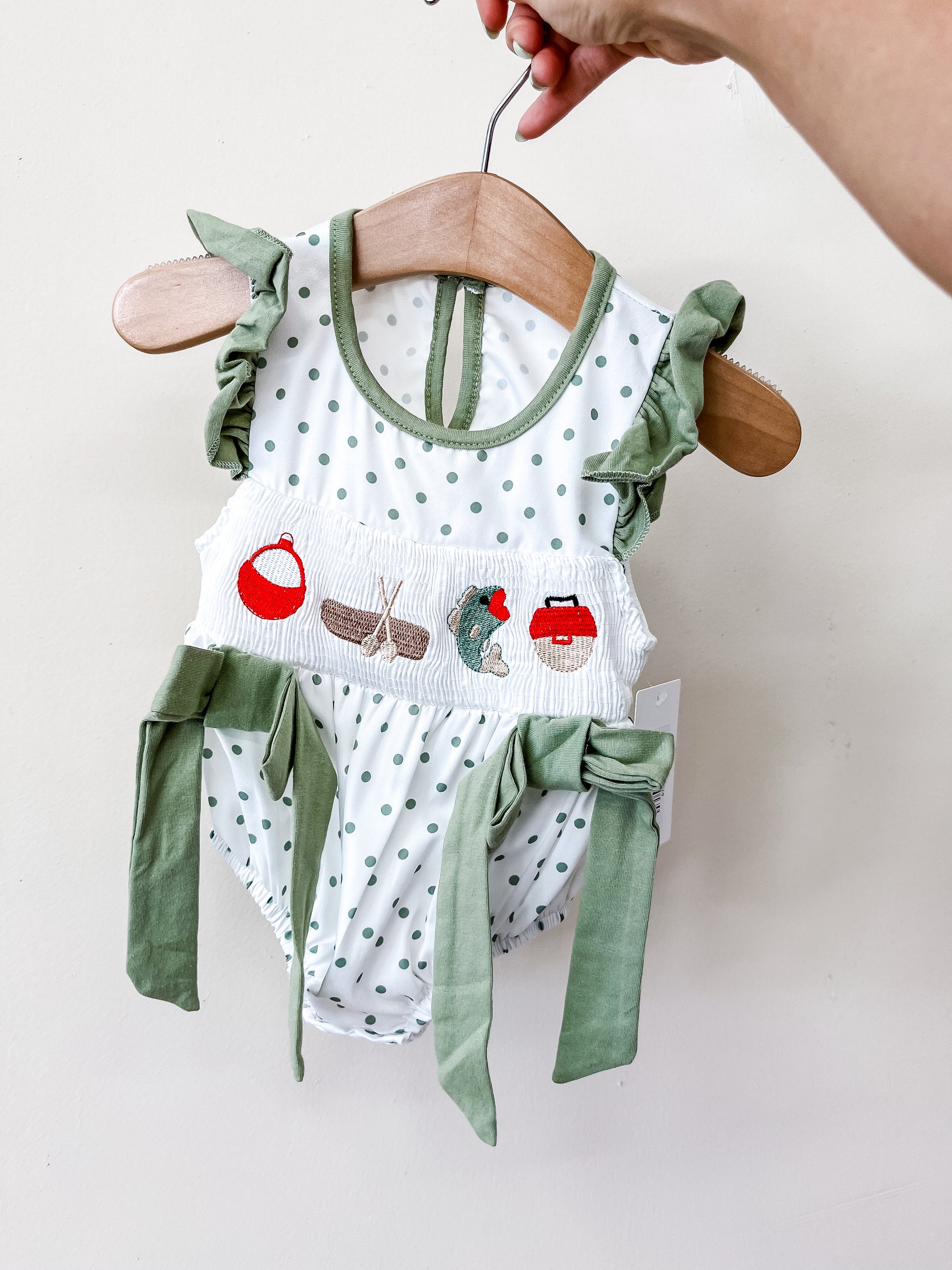 A hand holds up the Tiny Tot Bundles Polka Dot Pond Romper, featuring green and white polka dots, fishing-themed embroidery, ruffled sleeves, and side bows, displayed on a wooden hanger against a plain wall.