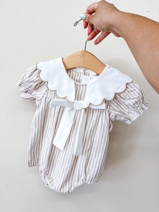 A hand holds a hanger displaying the Tiny Tot Bundles Coastal Classic Romper, featuring beige and white stripes, puff sleeves, a large scalloped white collar, and a front bow, against a plain white background.