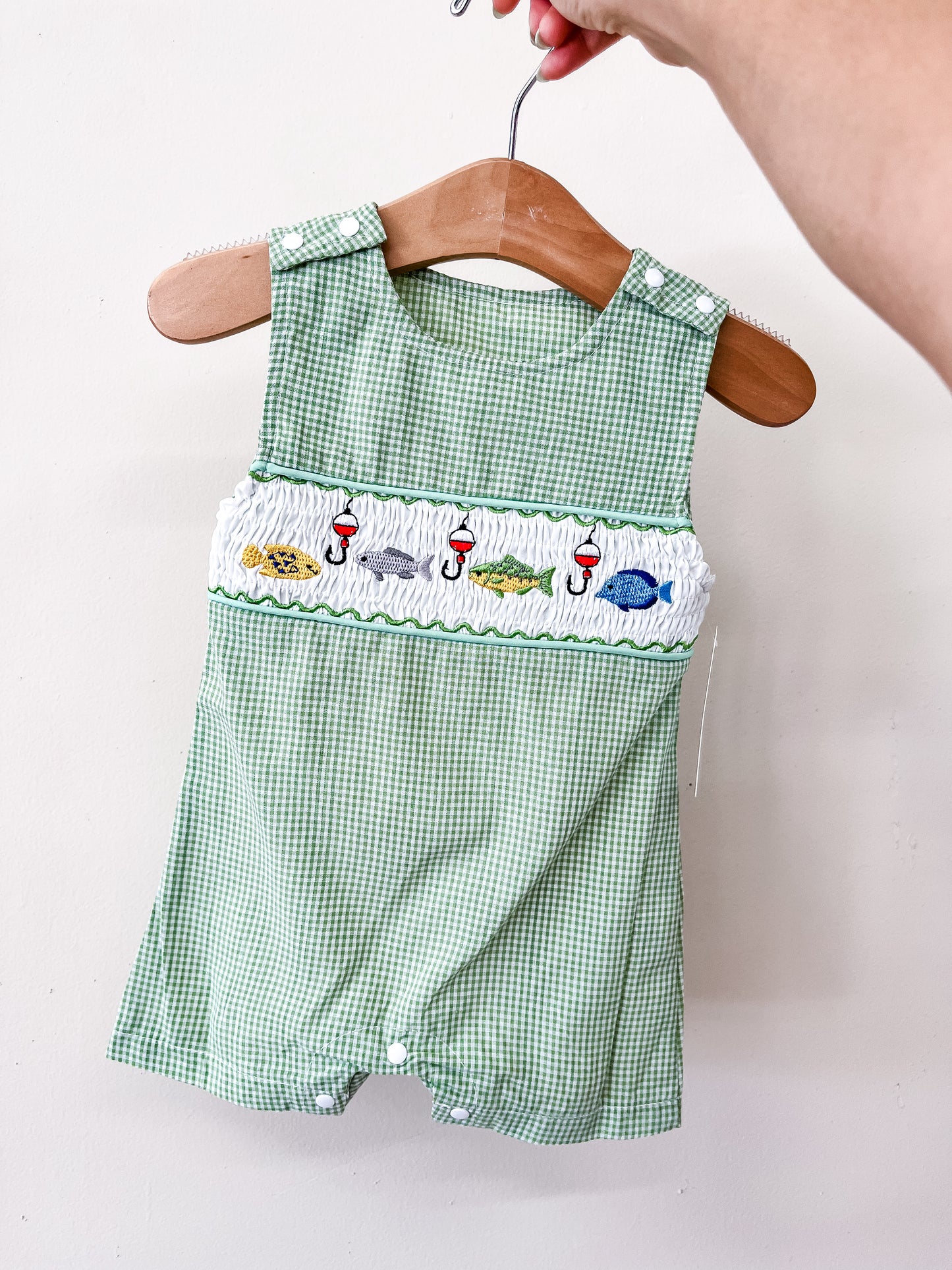 The Catch Of The Day Romper by Tiny Tot Bundles is displayed on a wooden hanger and features green checkered fabric with embroidered fish, sharks, and bobbers across the chest. A hand holds the hanger against a plain white background.
