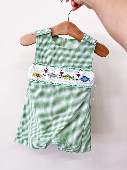 The Catch Of The Day Romper by Tiny Tot Bundles is displayed on a wooden hanger and features green checkered fabric with embroidered fish, sharks, and bobbers across the chest. A hand holds the hanger against a plain white background.