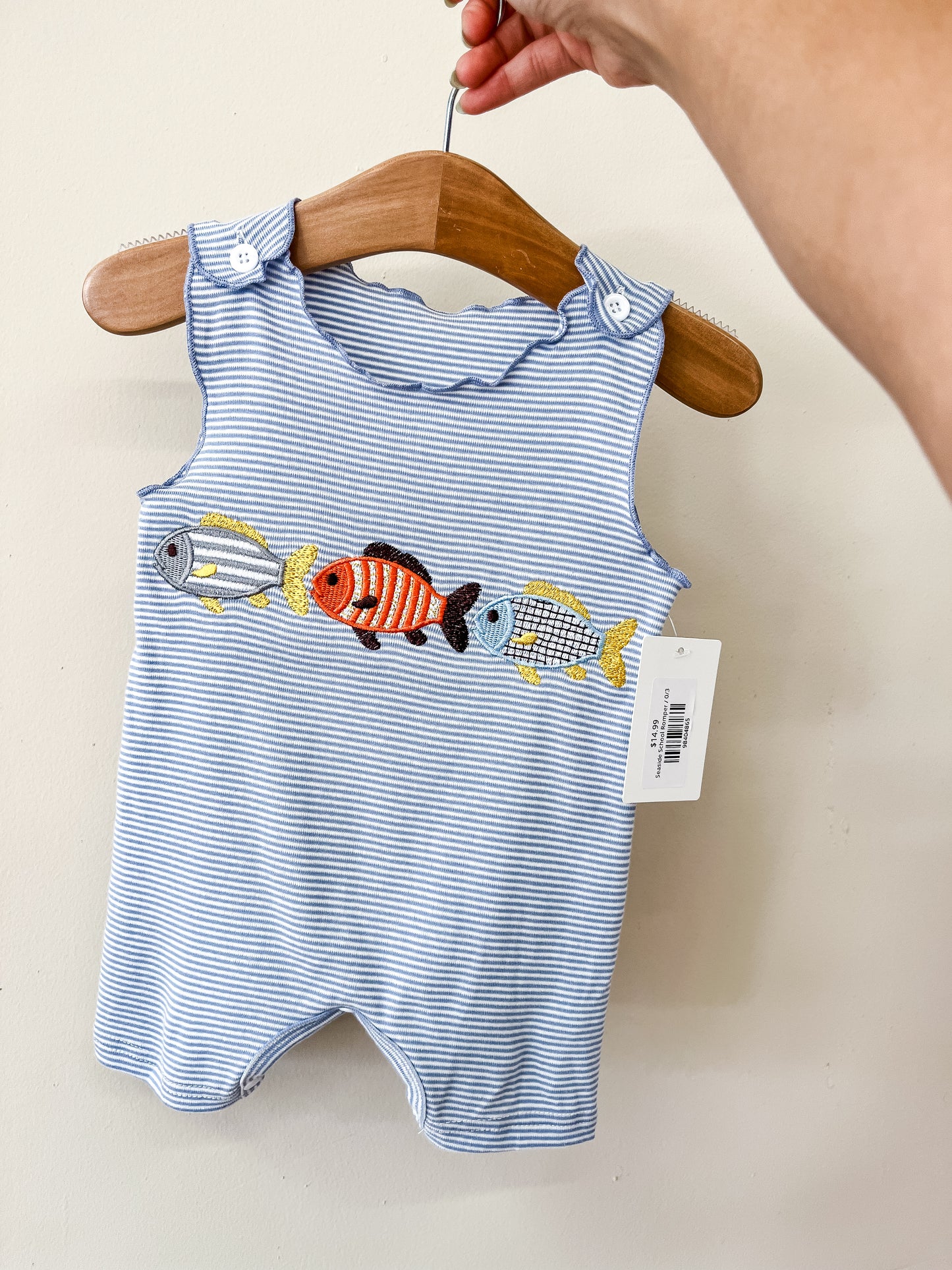 A hand holds a wooden hanger displaying the Tiny Tot Bundles Seaside School Romper—a blue and white striped baby romper with embroidered colorful fish across the chest. A price tag is attached.