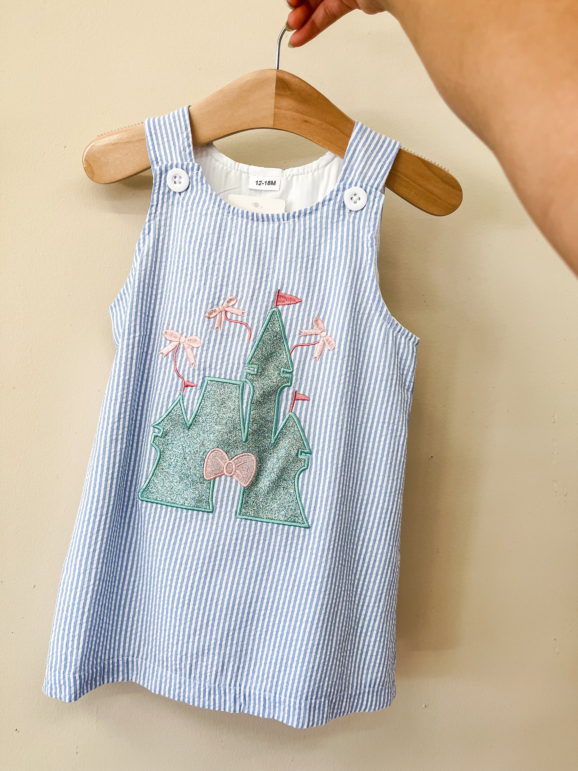 A hand holds a hanger with the Enchanted Castle Dress by Tiny Tot Bundles, a blue and white striped toddler dress featuring an embroidered castle with flags and a bow on the front.