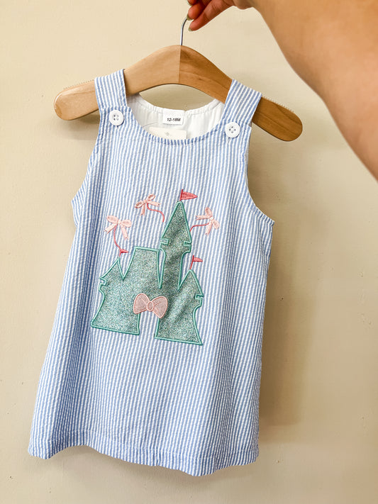 A hand holds a hanger with the Enchanted Castle Dress by Tiny Tot Bundles, a blue and white striped toddler dress featuring an embroidered castle with flags and a bow on the front.