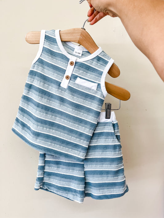 A hand holds a hanger displaying the Tiny Tot Bundles Blue Harbor Short Set, a matching blue and white striped toddler outfit with a sleeveless, button-front top featuring a pocket, paired with coordinating shorts.