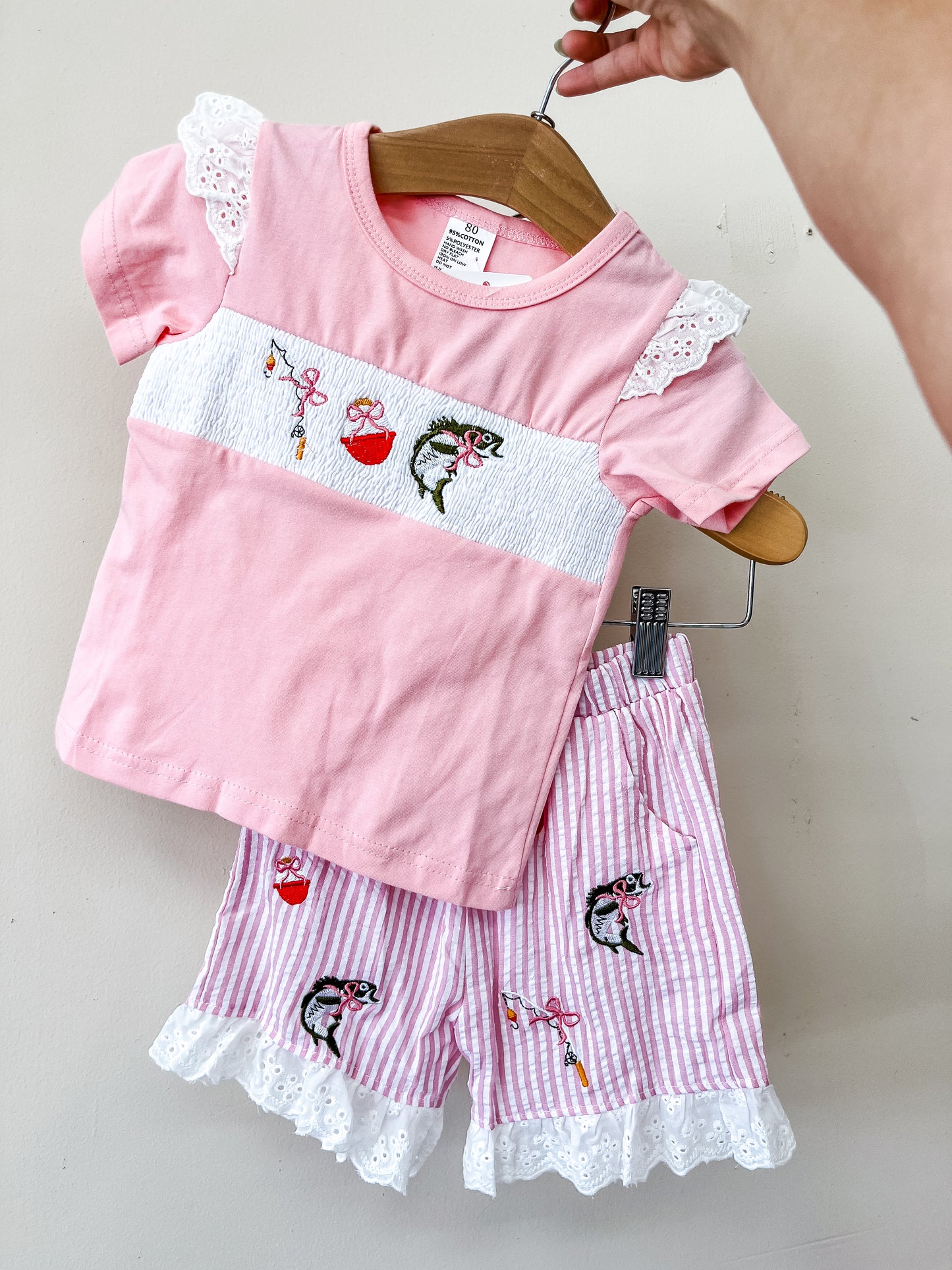 The Pretty Pink Fish Set by Tiny Tot Bundles features a lace-trimmed short-sleeve top and striped shorts, both adorned with charming embroidered cherries and birds. A hand displays the outfit on a wooden hanger against a plain off-white wall.