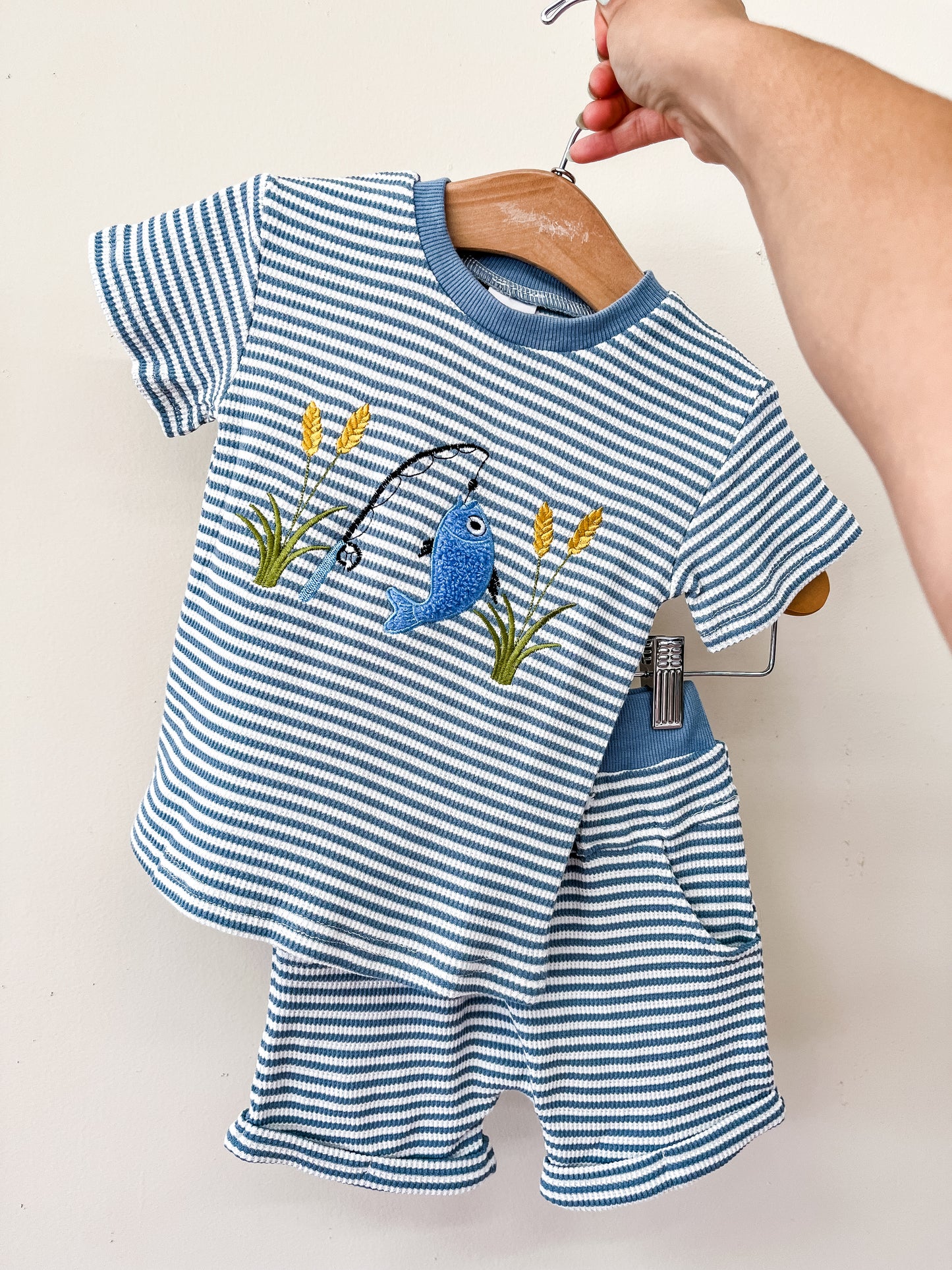 A hand holds a hanger displaying the "Hooked On Blue Set" by Tiny Tot Bundles, featuring a toddler short-sleeve shirt with embroidered fish, fishing pole, and grass, paired with matching blue and white striped shorts with a pocket.