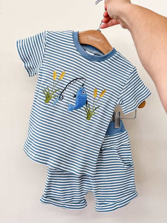 A hand holds a hanger displaying the "Hooked On Blue Set" by Tiny Tot Bundles, featuring a toddler short-sleeve shirt with embroidered fish, fishing pole, and grass, paired with matching blue and white striped shorts with a pocket.