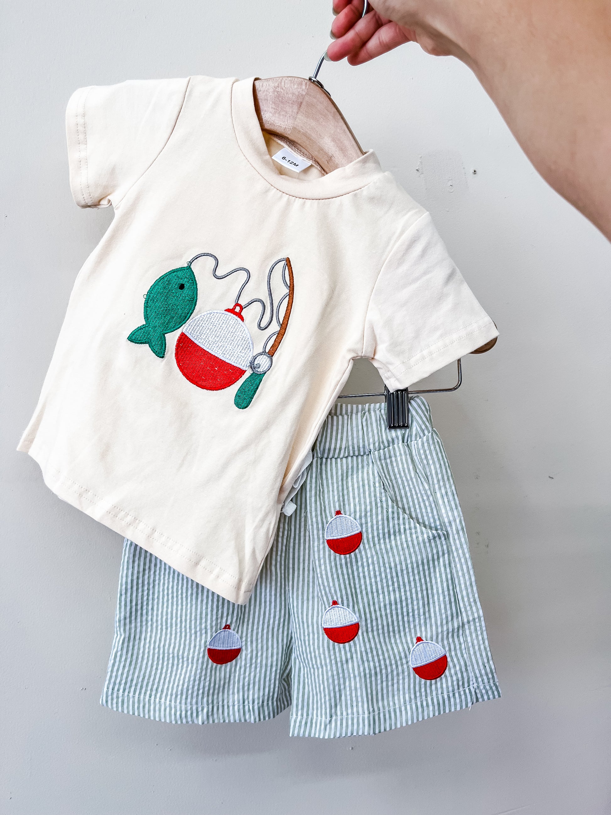 A hand holds up the Reel Adventure Short Set from Tiny Tot Bundles: a cream t-shirt featuring a fishing pole, fish, and bobber design, paired with light blue striped shorts adorned with red and white bobbers—all displayed on a hanger against a white background.
