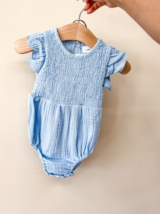The Blue Timeless Romper by Tiny Tot Bundles, a light blue, short-sleeved baby romper with ruffle details, is displayed on a wooden hanger held by a hand against a plain beige background.