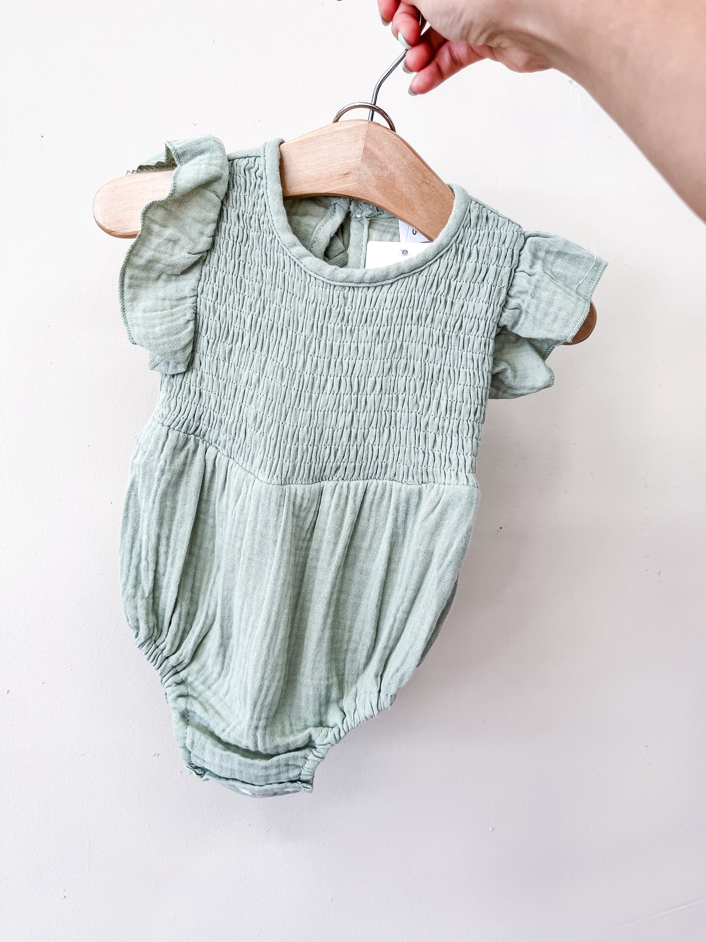 A hand holds a wooden hanger displaying the Tiny Tot Bundles Green Timeless Romper— a light green baby romper with short sleeves, ruffled shoulders, and a smocked bodice — set against a plain white background.