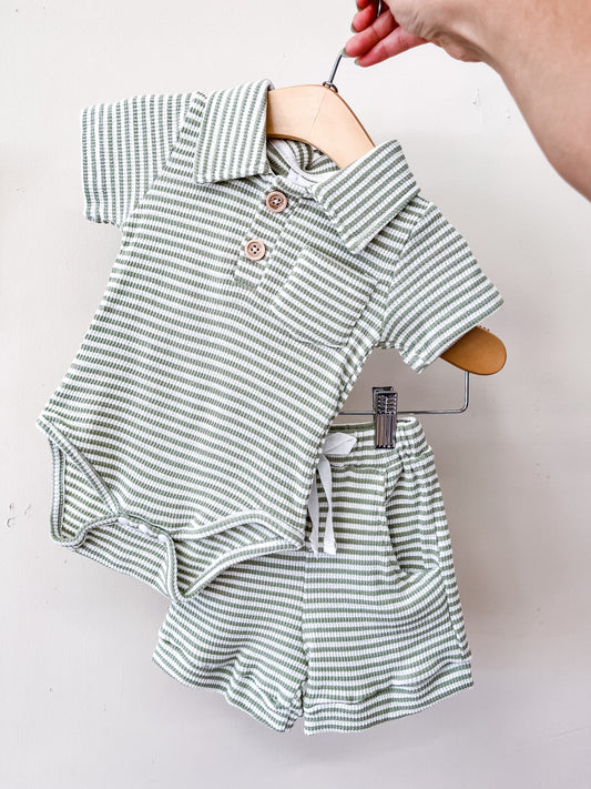 A hand holds a hanger displaying the Tiny Tot Bundles Green Harbor Polo Set, which features a green and white striped collared short-sleeve polo onesie with buttons paired with matching drawstring shorts.