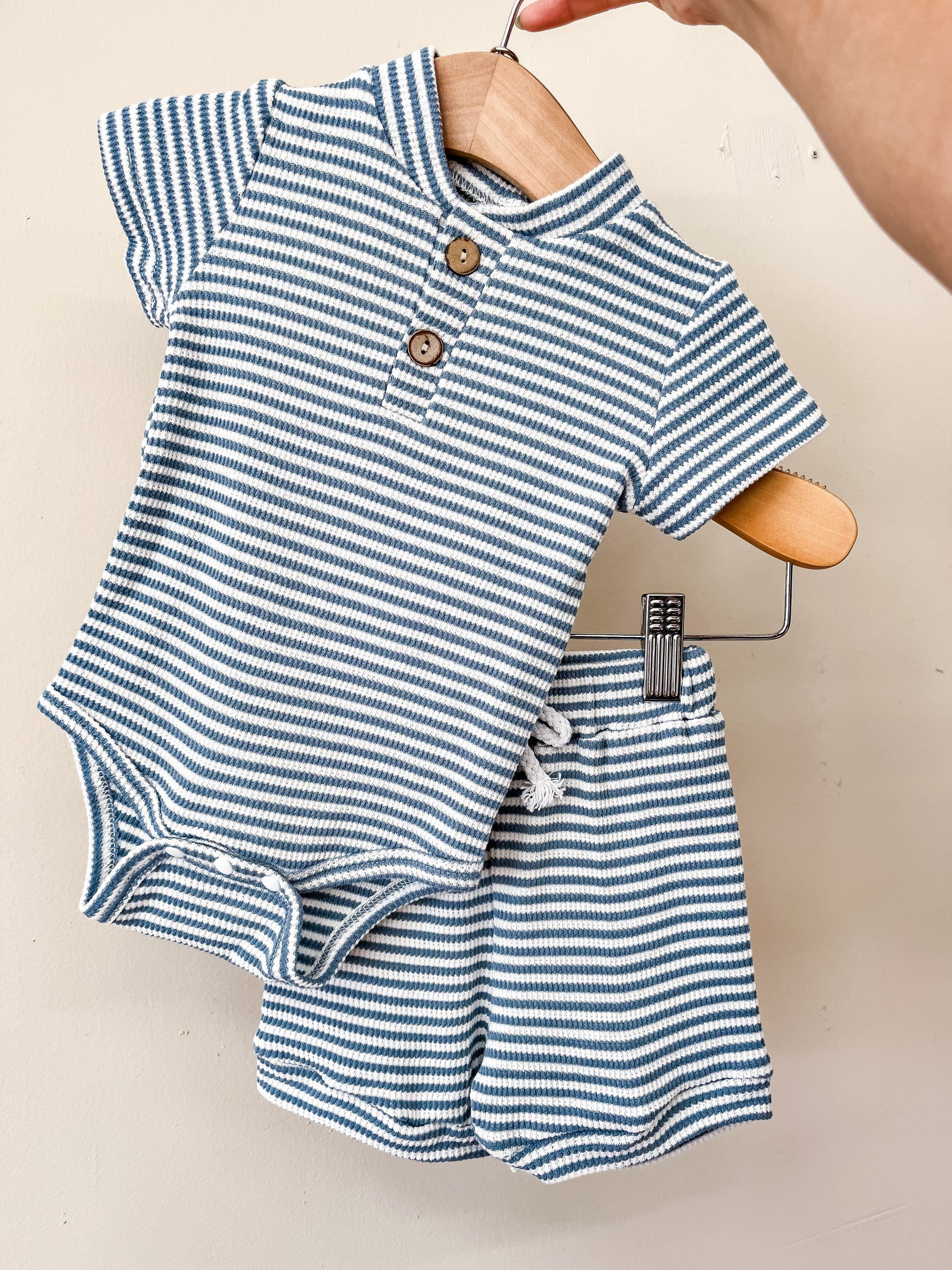 A hand holds a hanger displaying the Blue Harbor Polo Set by Tiny Tot Bundles, featuring a blue and white striped baby romper with brown buttons and matching ribbed shorts, set against a plain light background.