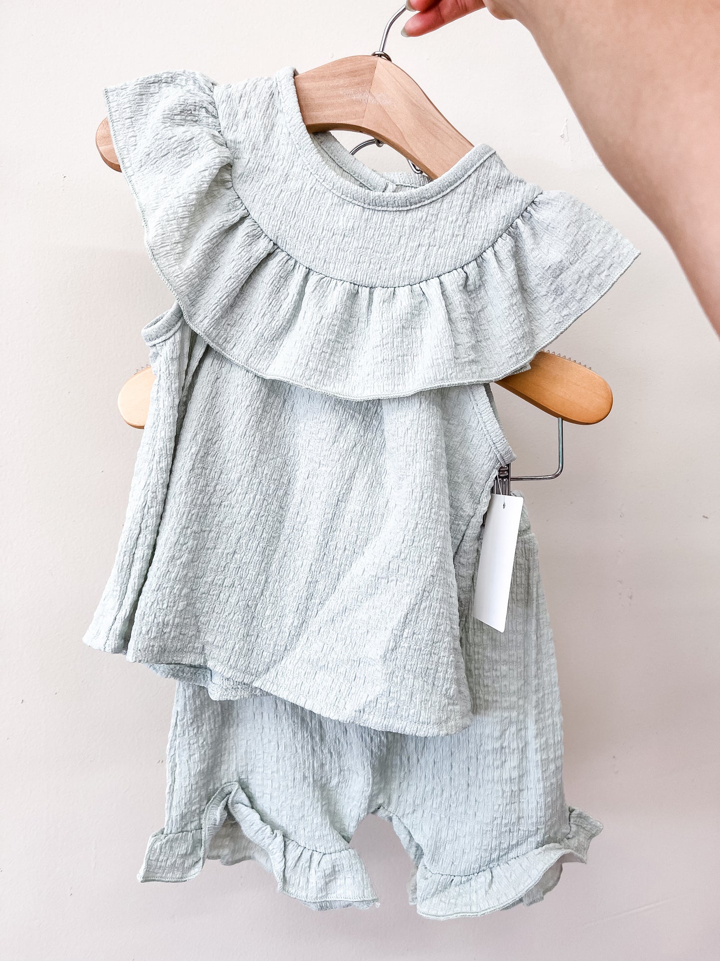 A hand holds a hanger with the Sage Meadow Short Set by Tiny Tot Bundles—a light gray, sleeveless baby outfit with a ruffled collar and matching textured shorts. A white tag hangs from the top.