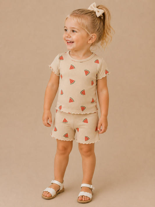 A young child stands and smiles in the Tiny Tot Bundles Watermelon Dreams Set—featuring a beige outfit with watermelon prints—paired with white sandals and a large bow, against a plain beige background.