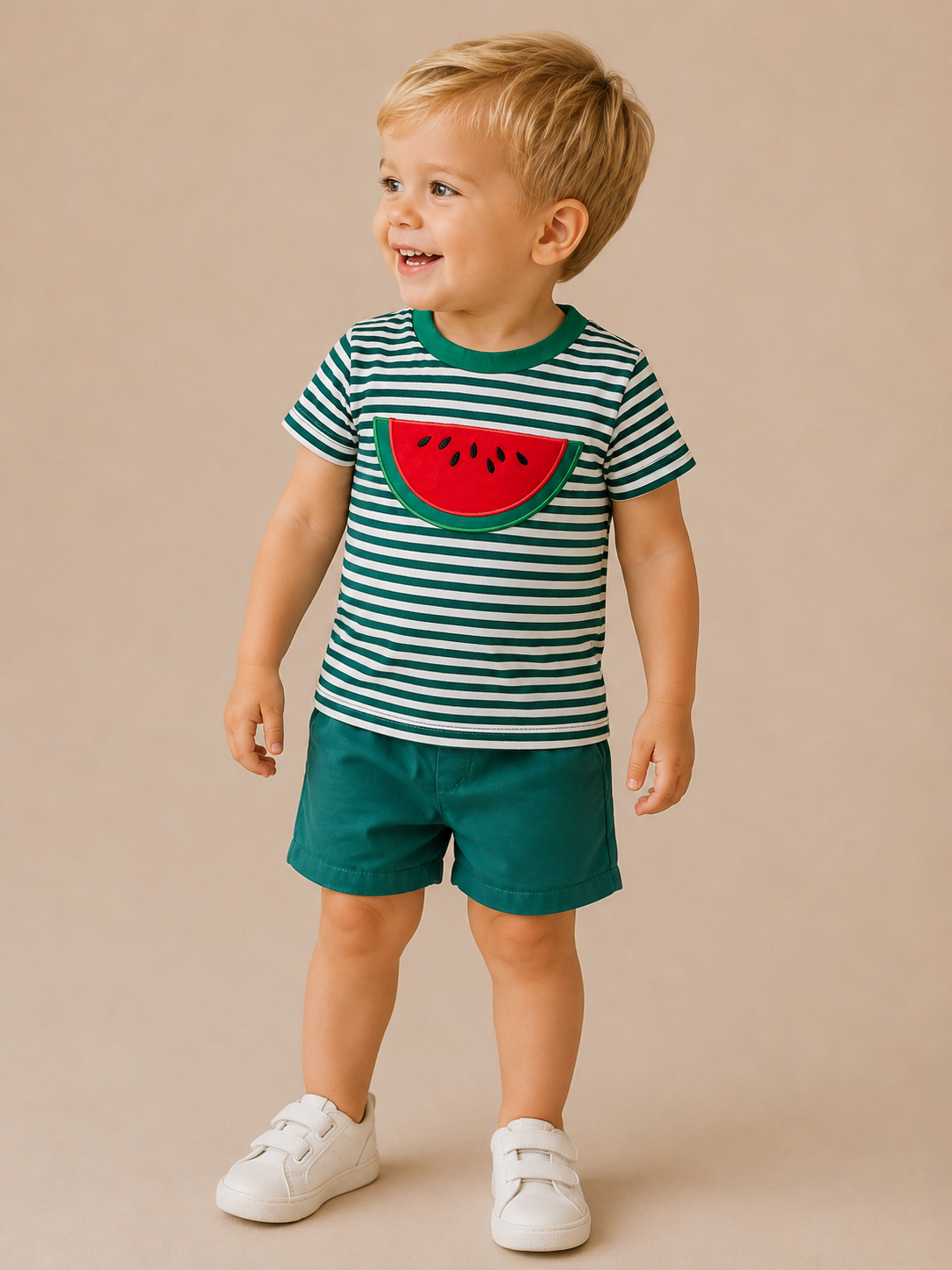 A smiling young child stands against a beige background, wearing Tiny Tot Bundles' Watermelon Adventure Set—featuring a green and white striped shirt with a watermelon graphic, dark green shorts, and white sneakers.
