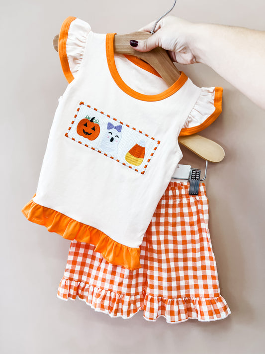The Spooky Sweet Gingham Set by Tiny Tot Bundles features a small white top with ruffled orange trim and Halloween-themed embroidery, paired with orange and white gingham ruffled shorts, displayed on hangers held by hand.