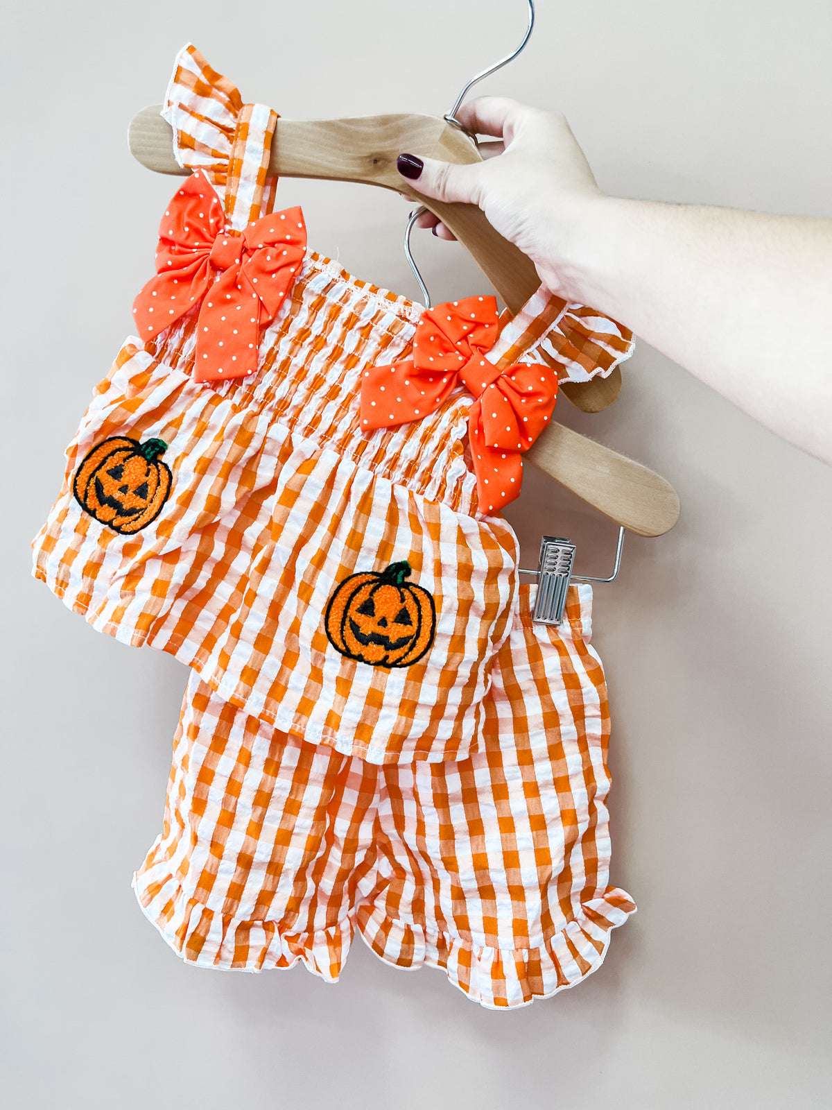 A hand holds the Pumpkin Patch Gingham Set by Tiny Tot Bundles—an orange and white gingham Halloween outfit featuring pumpkin patches and orange bow accents—displayed on a wooden hanger against a plain background.