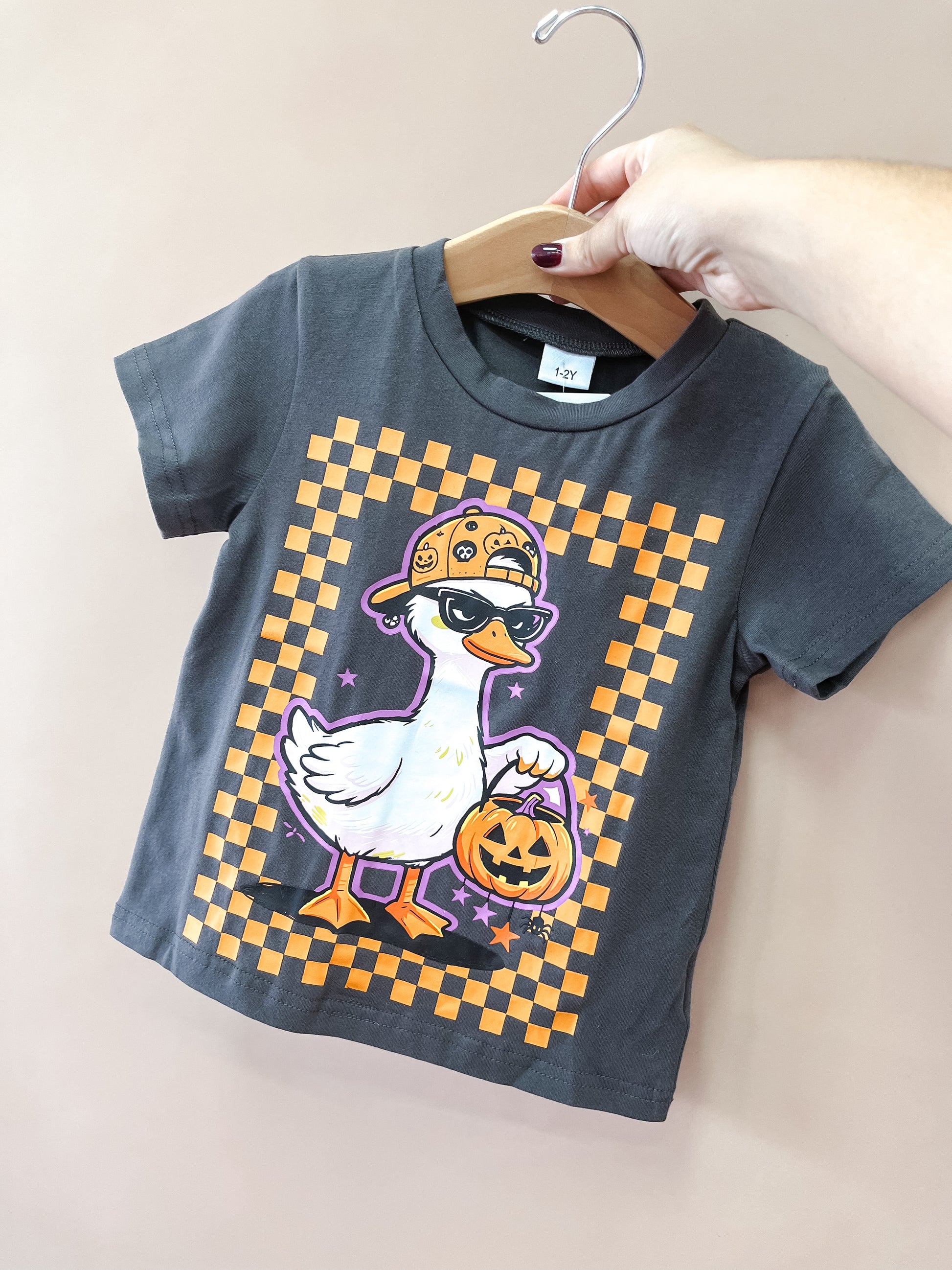 A hand holds up the Black Spooky Goose Tee by Tiny Tot Bundles, a gray children’s t-shirt featuring a cartoon goose in a cap and mask holding a pumpkin bucket, with an orange checkerboard border.