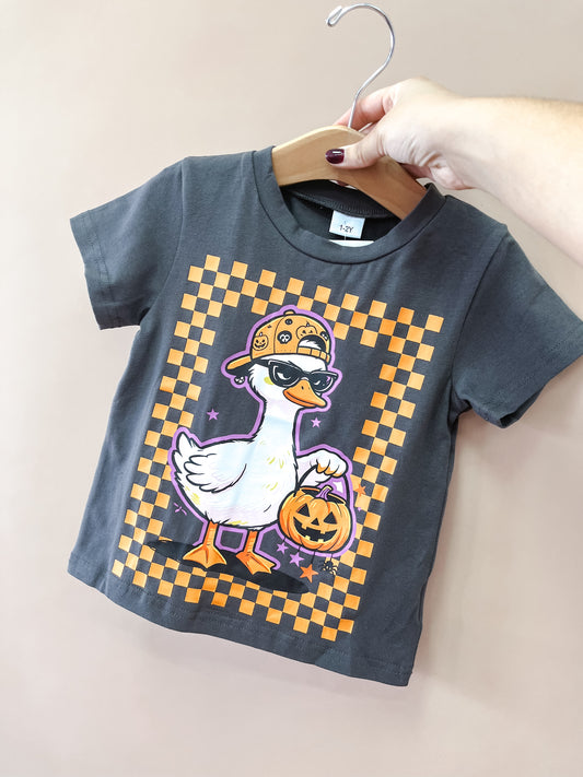 A hand holds up the Black Spooky Goose Tee by Tiny Tot Bundles, a gray children’s t-shirt featuring a cartoon goose in a cap and mask holding a pumpkin bucket, with an orange checkerboard border.
