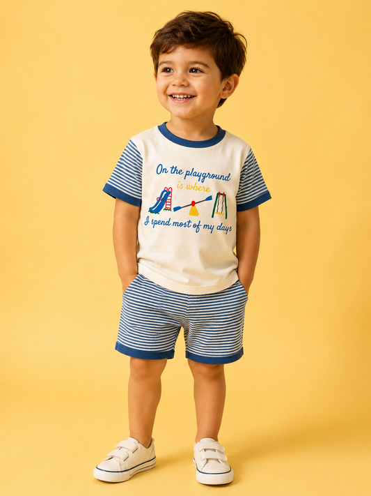 A smiling young boy wears the Playground Day's Set from Tiny Tot Bundles—featuring a cream and blue playground-themed t-shirt with blue and white striped shorts—posing with hands in pockets against a yellow background.