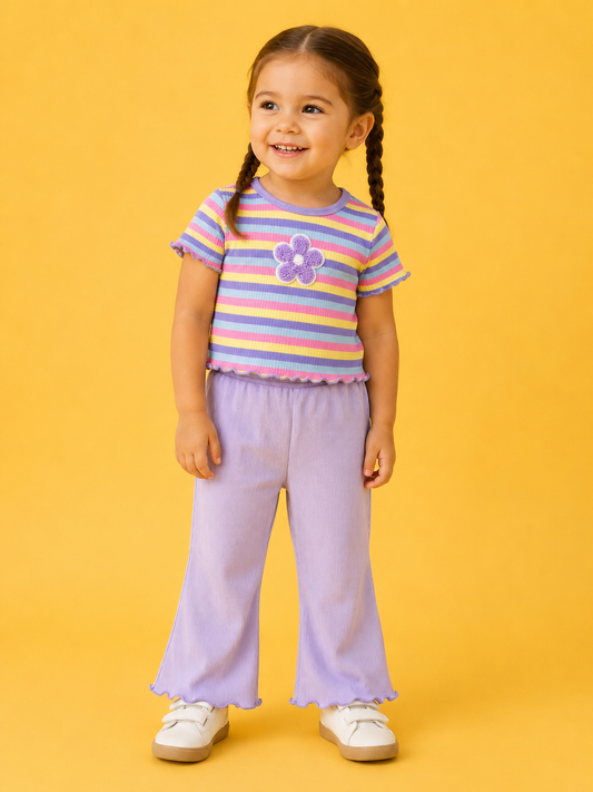 A young girl with brown braided hair smiles against a yellow background, wearing the Floral Rainbow Row Set from Tiny Tot Bundles: a striped pastel shirt with a purple flower, light purple pants, and white shoes.