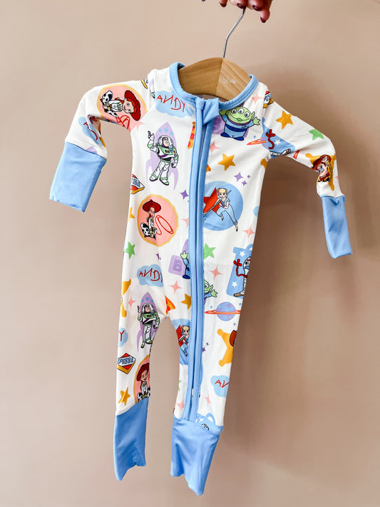 Someone holds the Toy Box Pals Zippy by Tiny Tot Bundles, a baby onesie with blue cuffs and zipper, decorated with Toy Story characters and colorful stars on a white background.