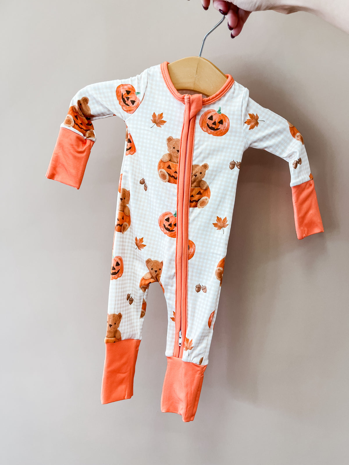 Tiny Tot Bundles Pumpkin Bear Zippy long-sleeved baby sleeper featuring a pattern of teddy bears, pumpkins, and autumn leaves on a white checkered background with solid orange cuffs.