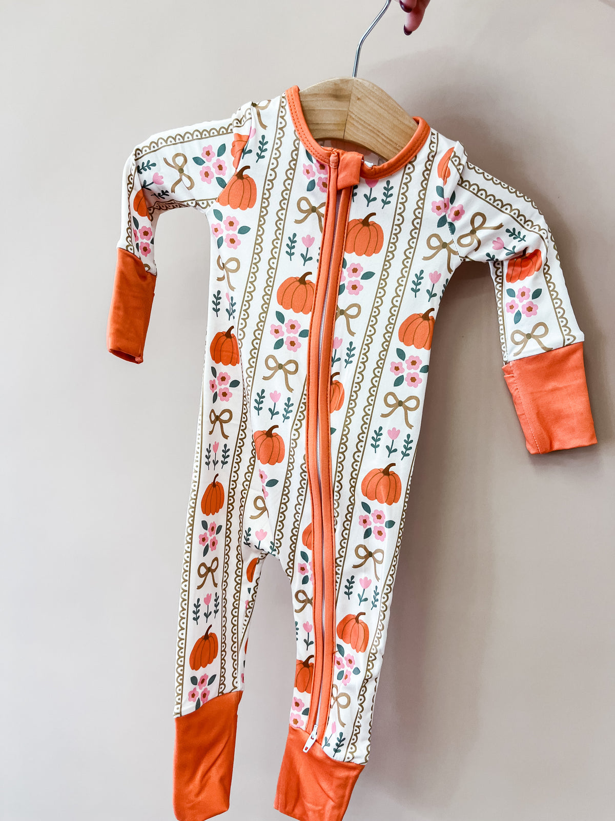 Tiny Tot Bundles Autumn Bows Zippy, a long-sleeved baby zip-up sleeper featuring a pattern of pumpkins, floral vines, and bows with orange trim on the collar, cuffs, and ankles.