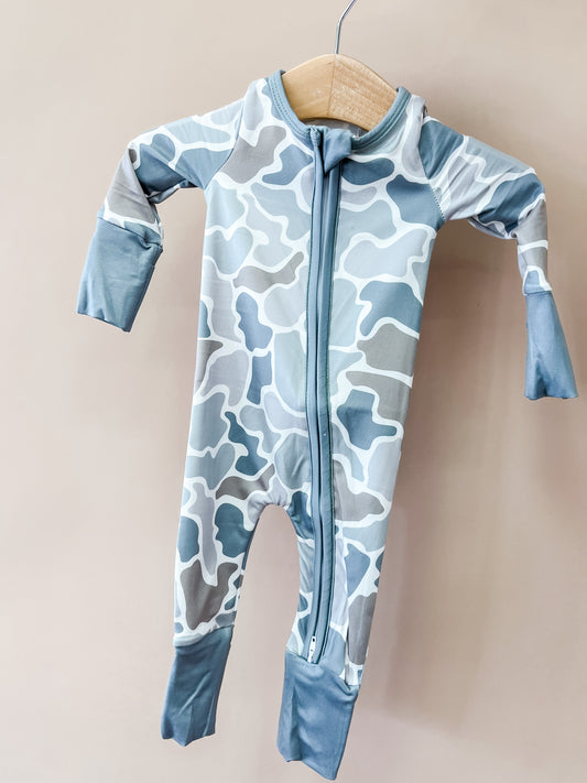 A Wild Country Zippy baby onesie by Tiny Tot Bundles, featuring a blue and gray camouflage pattern with long sleeves and a front zipper, hangs on a wooden hanger against a beige background.