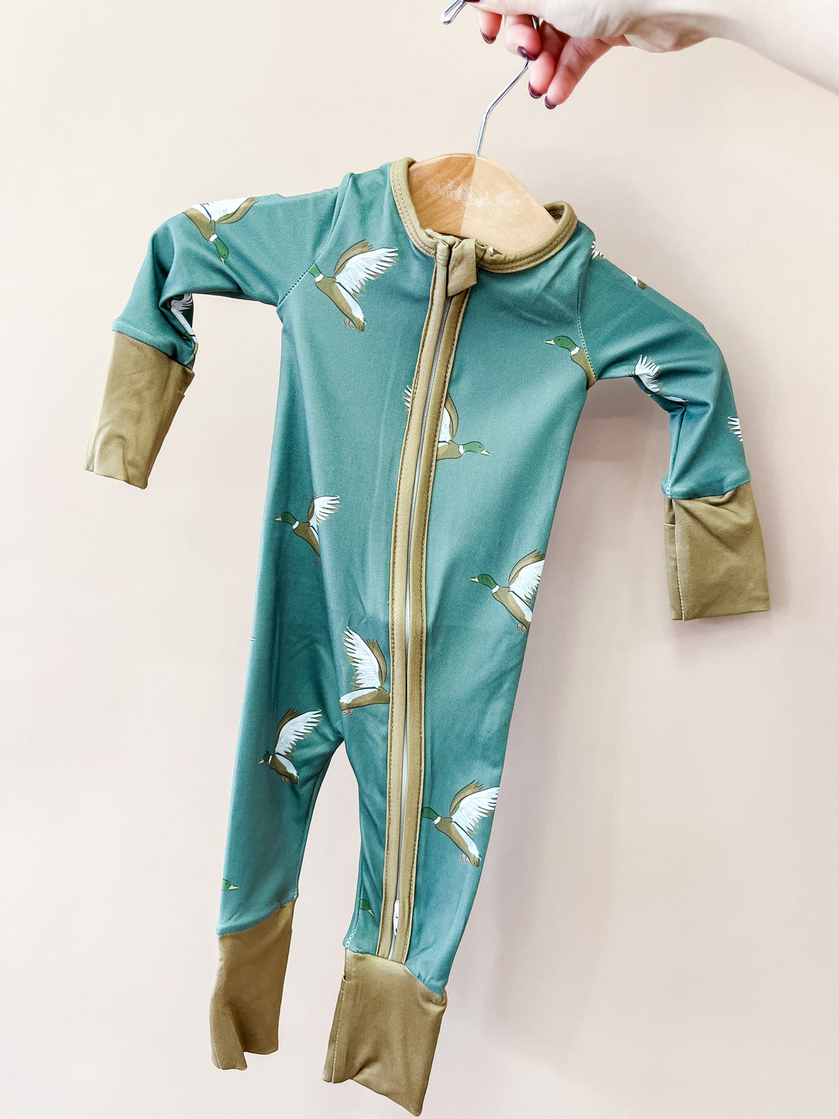 A hand holds a hanger with the Tiny Tot Bundles Evergreen Mallard Zippy—a green baby onesie adorned with flying duck prints, tan cuffs, and a front zipper—set against a beige background.