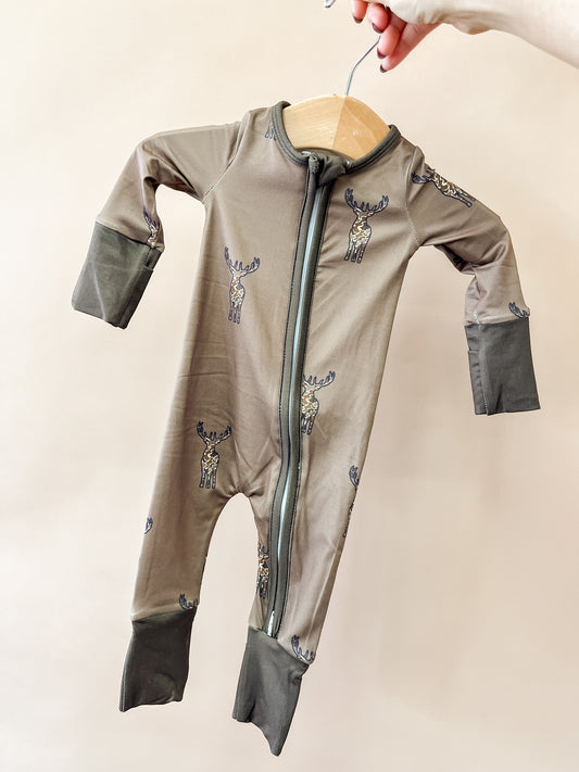 A hand holds a hanger with the Tiny Tot Bundles Wild Woods Zippy, a brown baby onesie adorned with moose prints, dark cuffs, and a front zipper, set against a plain light background.