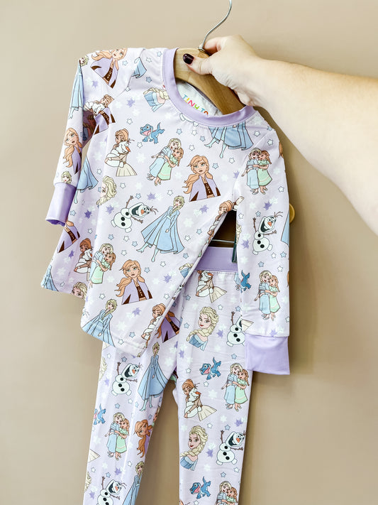 A hand holds up the Snow Sisters Jammies Set by Tiny Tot Bundles, featuring Anna, Elsa, Olaf, and snowflakes on a purple background.