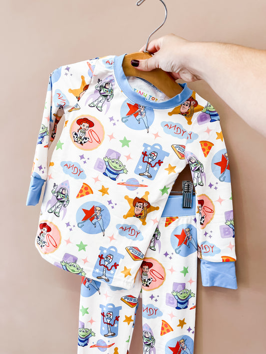 Hand holding a hanger with Tiny Tot Bundles Toy Box Pals Jammies Set—children’s pajamas featuring Woody, Buzz Lightyear, and aliens on a pizza, stars, and blocks background—against a plain wall.